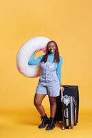 africano americano niña dejando en fiesta con inflable anillo y carretilla bolsas, sensación emocionado acerca de vacaciones. joven mujer disfrutando fin de semana ocupaciones en el extranjero, que lleva equipaje. foto