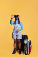 Young adult having fun with vr glasses in studio, posing over orange background with virtual reality headset and trolley bags. Female traveller using modern goggles with 3d vision. photo