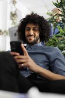 Smiling arab executive manager texting in social network on smartphone in business office. Start up company employee chatting on mobile phone while sitting on couch in coworking space photo