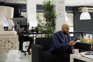 Smiling business entrepreneur answering text message on smartphone in start up office. Young arab businessman using social media on mobile phone while having break at work photo