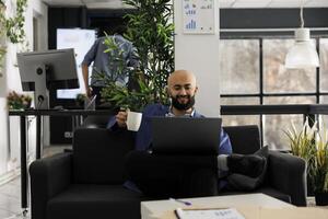 Smiling relaxed startup business employee drinking coffee and working on laptop in office. Happy arab man sitting on couch and holding tea mug while checking successful marketing results photo