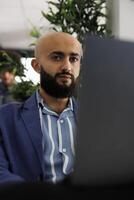 Arab businessman sitting in front of laptop at desk, preparing for video call with clients. Executive manager checking financial analytics report before presentation in coworking space photo