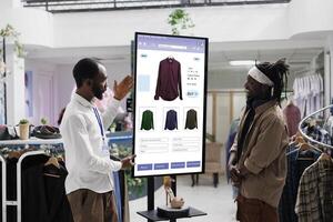 Young worker showing items on touch screen monitor kiosk service to client in fashion boutique at mall. Store assistant advising customer to select trendy clothing line merchandise. photo