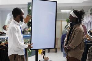 African american employee using smart display to assist customer with shoe purchase in shopping center. Clothing store showing promotion on whiteboard, helping customers selecting stiletto photo