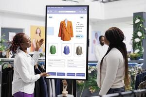 People looking at interactive board clothing store kiosk used for self ordering service at mall, customer choosing trendy clothes to buy from touch screen monitor board. E commerce concept. photo