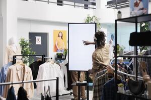 africano americano hombre mientras utilizando inteligente interactivo monitor tablero en ropa almacenar. cliente tocando en blanco blanco pantalla burlarse de arriba para Zapatos digital anuncios y información en compras centro comercial boutique foto