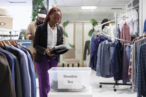 compras centro comercial africano americano cliente poniendo ropa en donación caja a ayuda pobre. mujer contribuyendo a caridad trabajar como voluntario organización, dando vestir para humanitario ayuda foto