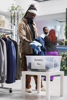 africano americano hombre donando antiguo ropa a caridad organización en compras centro comercial. Moda boutique cliente poniendo formal ropa en envase para bienestar a ayuda pobre foto