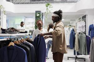 African american buyer and seller discussing fashion trends while choosing jacket in clothing store. Shopping mall customer and assistant browsing through rack with hanging blazers photo