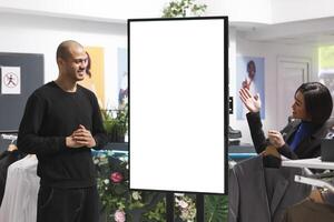 Clothing store smiling asian woman employee pointing at empty interactive board while giving client advice. Boutique consultant holding and showing customer advertising on blank whiteboard photo