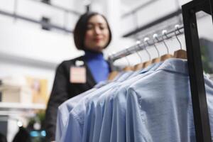 formal camisas colgando en monitor estante mientras asiático mujer asistente trabajando en ropa Tienda selectivo enfocar. ropa de caballero prenda en perchas en compras centro comercial boutique de cerca foto