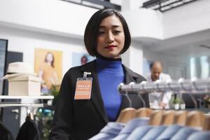 Asian woman seller working in clothing store, managing formal apparel inventory and checking shits on display rack. Shopping center employee organizing menswear garment in boutique photo