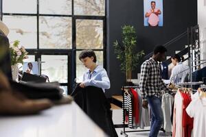 Store worker holding stylish shirts, putting fashionable merchandise on hangers in shopping centre. Cheerful manager arranging casual wear, checking clothes price in modern boutique photo