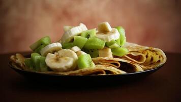 cooked thin pancakes with bananas and kiwi on a light wooden table video