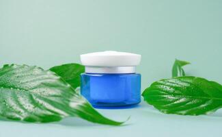 Blue glass jar with white lid with face cream with green leaves, green background, copy space. spa product. Organic, bio, natural cosmetics. Beauty, skin care concept. photo