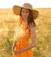 Young woman in an orange dress and a straw hat standing on a field in the rays of the setting sun photo