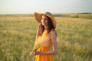 Young woman in an orange dress and a straw hat standing on a field in the rays of the setting sun photo