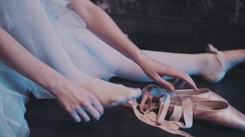 A ballerina in a classic white suit puts on pointe shoes while sitting on stage before a performance. Close-up video