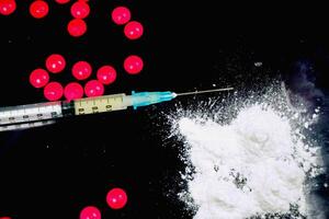 Top view and crop of plastic syringe in a medical needle and white powder with red pill medicine isolate on black background. Suppose to heroin and amphetamine or narcotic addiction. photo