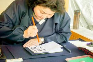 Kioto ciudad, Japón, 2023 - japonés mujer vistiendo un azul nacional vestir y participación un chino cepillo escritura japonés textos desde negro tinta en un cuaderno papel. foto