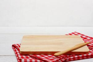 Empty rectangle wooden serving board with butter knife and red gingham tablecloth on white wooden table. photo