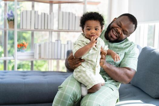 contento africano americano hombre jugar con pequeño infantil niñito niño en vivo habitación sofá. padre y pequeño niño relajante a hogar. padres y niñito niño niña disfrutando gasto hora a hogar. cuidado de niños foto
