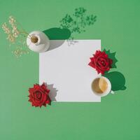 Creative spring layout made with cup of coffee, red roses, white vase with dry flowers and paper card note copy space on green background. Minimal concept. Trendy coffee with flowers idea. Flat lay. photo