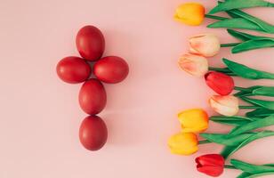 Creative Easter holiday composition of red Easter eggs in a shape of a cross and colorful tulips on pastel pink background. Minimal concept. Easter eggs with spring flowers idea. Nature flat lay. photo