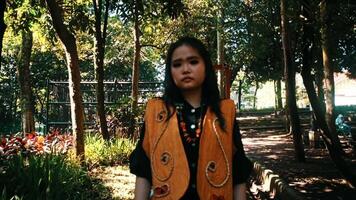 Serious young woman in a vibrant orange vest standing in a shaded park with sunlit trees video