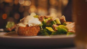 Close-up of a Caesar salad with croutons and dressing on a plate, with a blurred restaurant video