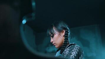 Portrait of a woman with braided hair, wearing armor, playing a grand piano and looking contemplative against a dark background. video