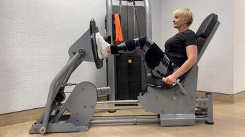 excesso de peso mulher 55-60 anos velho dentro a academia. uma mulher dentro a Academia faz perna pressione em a máquina. video