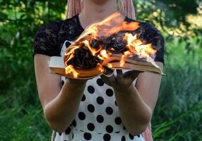 girl holds a burning book in her hands. A young woman in a forest burns a book. photo