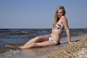 el niña el rubia en un bikini sentado en el playa en el arena. hermosa joven mujer en un vistoso bikini en mar antecedentes foto