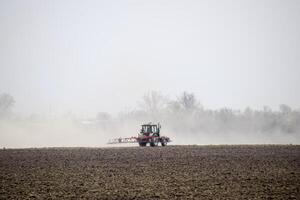 The tractor harrows the soil on the field and creates a cloud of dust behind it photo