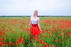 Blonde young woman in red skirt and white shirt, red earrings is in the middle of a poppy field. photo