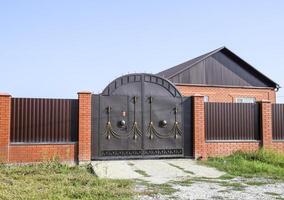 Brick house with a fence and gates. View of a new built-up fence and a house made of bricks and corrugated metal. photo