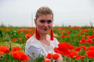 Blonde young woman in red skirt and white shirt, red earrings is in the middle of a poppy field. photo