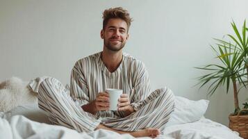 ai generado joven hermoso hombre en a rayas pijama bebidas café mientras sentado en el cama foto