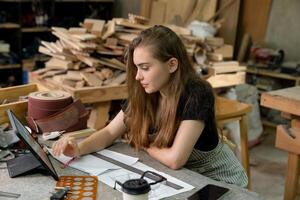 un joven mujer es formación a ser un carpintero en el taller. ella trabajos con un ordenador portátil computadora en un madera taller. hembra carpintero contacto clientes por teléfono inteligente SME pedidos, puesta en marcha y pequeño foto