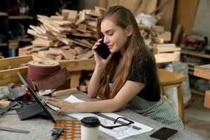 un joven mujer es formación a ser un carpintero en el taller. ella trabajos con un ordenador portátil computadora en un madera taller. hembra carpintero contacto clientes por teléfono inteligente SME pedidos, puesta en marcha y pequeño foto