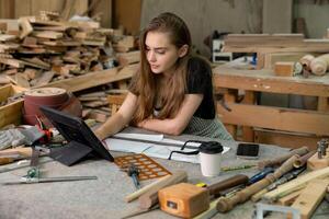 A young woman is training to be a carpenter in the workshop. She works with a laptop computer in a wood workshop. female carpenter contact customers by smartphone. SME orders, Start-up and small photo