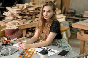 A young woman is training to be a carpenter in the workshop. She works with a laptop computer in a wood workshop. female carpenter contact customers by smartphone. SME orders, Start-up and small photo