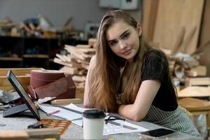 A young woman is training to be a carpenter in the workshop. She works with a laptop computer in a wood workshop. female carpenter contact customers by smartphone. SME orders, Start-up and small photo