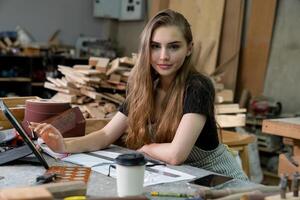 A young woman is training to be a carpenter in the workshop. She works with a laptop computer in a wood workshop. female carpenter contact customers by smartphone. SME orders, Start-up and small photo