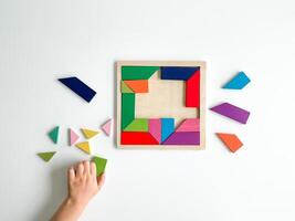 childs hand collects multicolored wooden mosaic on white background. child solves colorful tangram photo