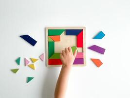 childs hand collects multicolored wooden mosaic on white background. child solves colorful tangram photo