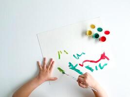 small child draws with paints and brush on white table. photo