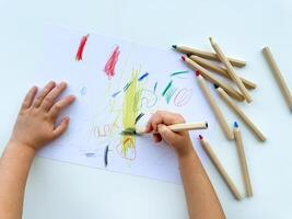 small child draws with colored pencils on paper on white table. photo