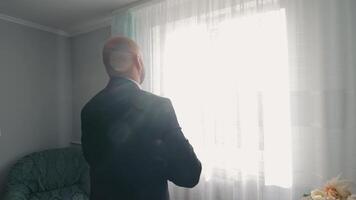 contemplativo hombre mirando mediante escarpado cortinas, un calvo hombre en un oscuro camisa mira fuera un brillante ventana cubierto con escarpado cortinas video